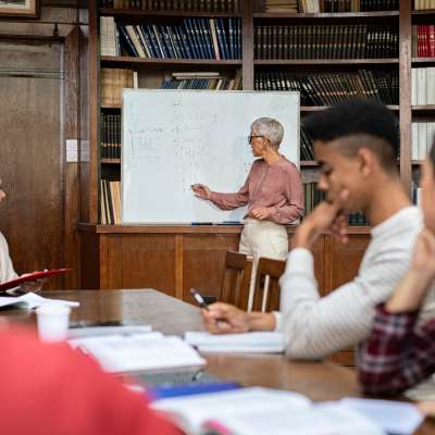 Senior maths teacher explaining math to a group of student using white board in library. Group of multiethnic university students understanding concept with helpful mature professor. Teacher teaching a class of guys and girls in library.